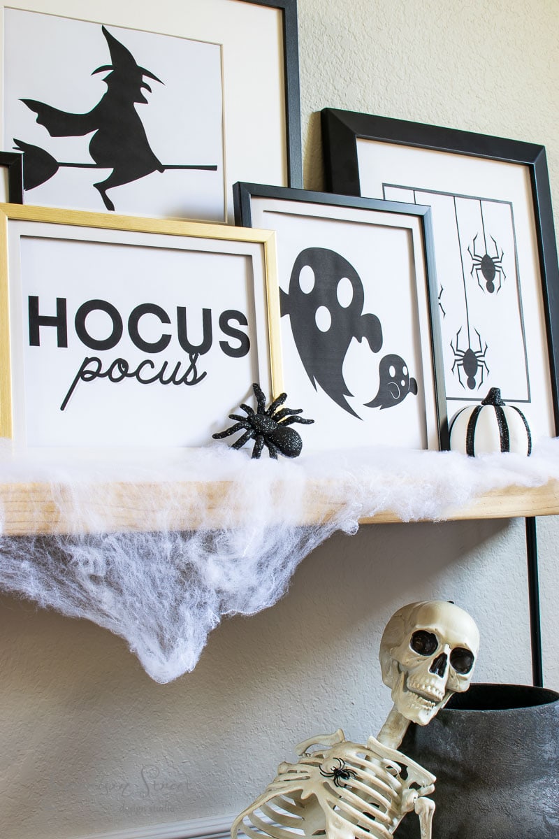 A shelf decorated for Halloween features framed black and white prints of witches, ghosts, and spiders. A fake spider and cobwebs sit on the shelf, with a skeleton posed in front.