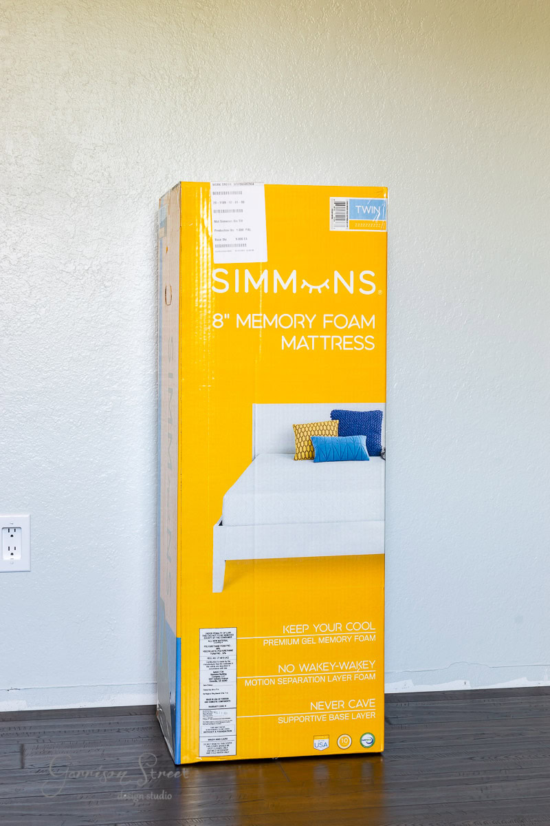A yellow cardboard box stands upright against a plain wall on a hardwood floor. The box, branded "Simmons," indicates it contains an 8" memory foam mattress, with illustrations and product features detailed on the front.