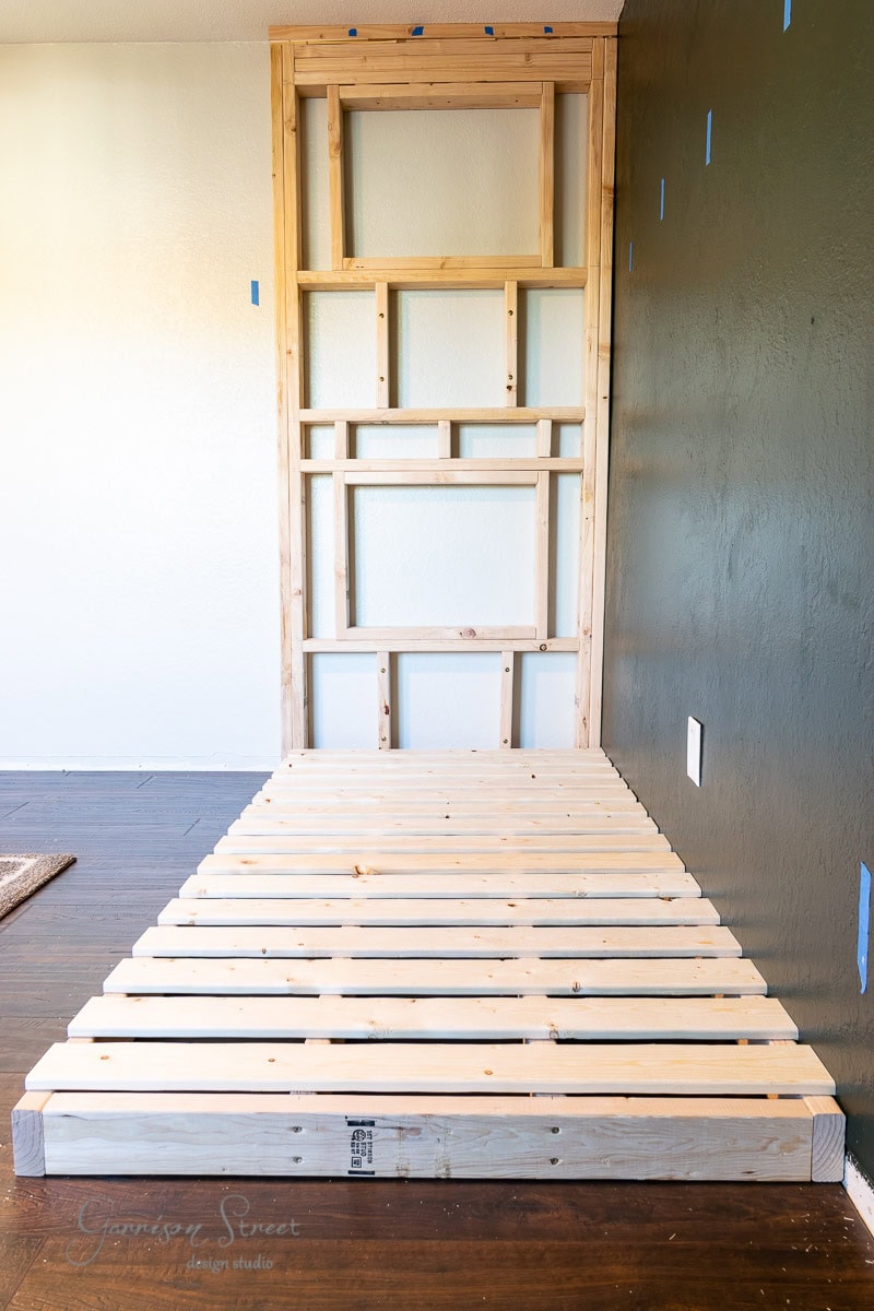 A wooden frame and slatted platform constructed against a wall in a room with dark hardwood floors and a dark green accent wall.