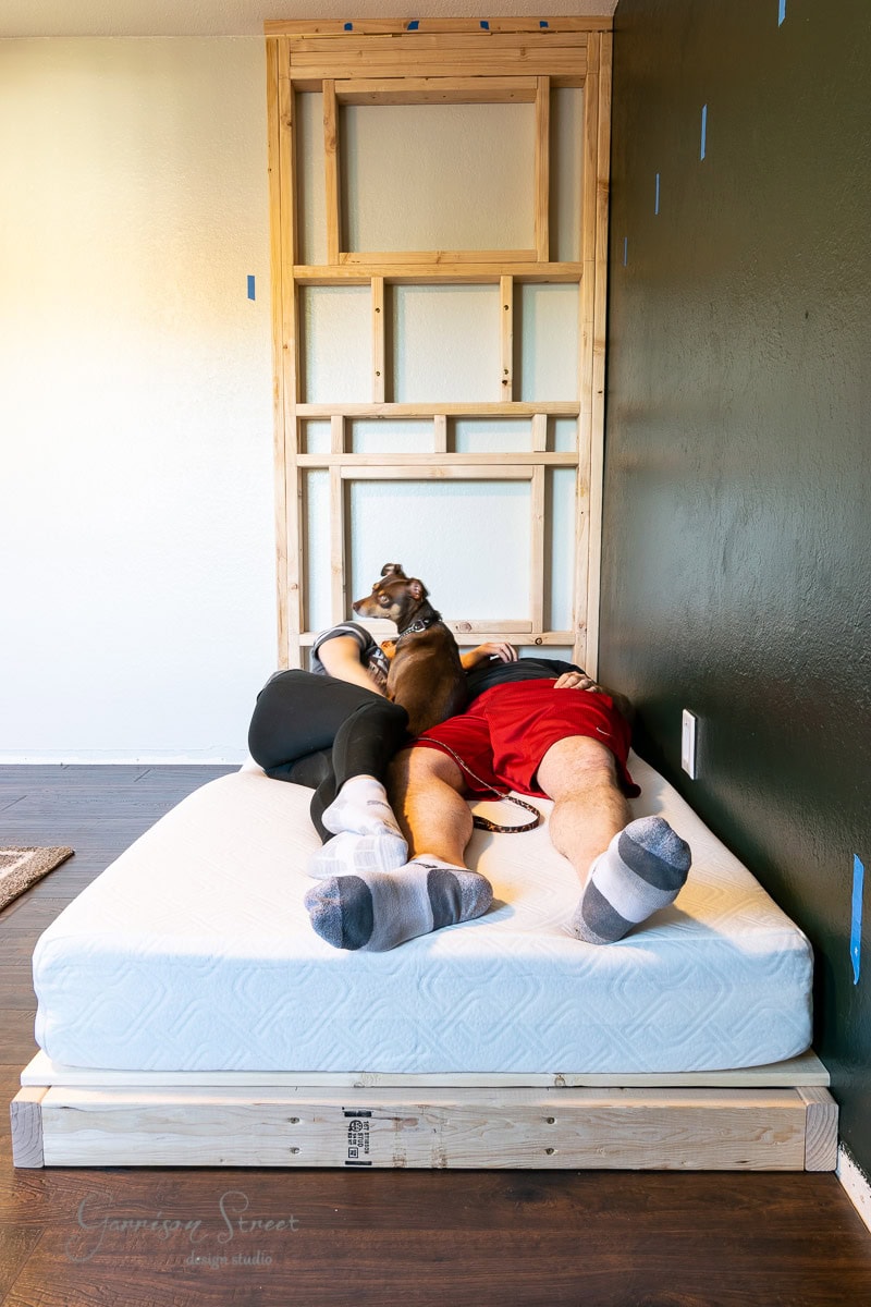 Two people lie on a mattress next to an unfinished wooden frame against the wall. They are wearing casual clothes and socks, with a small dog resting between them. The wall is partially painted dark, with blue tape marking sections.
