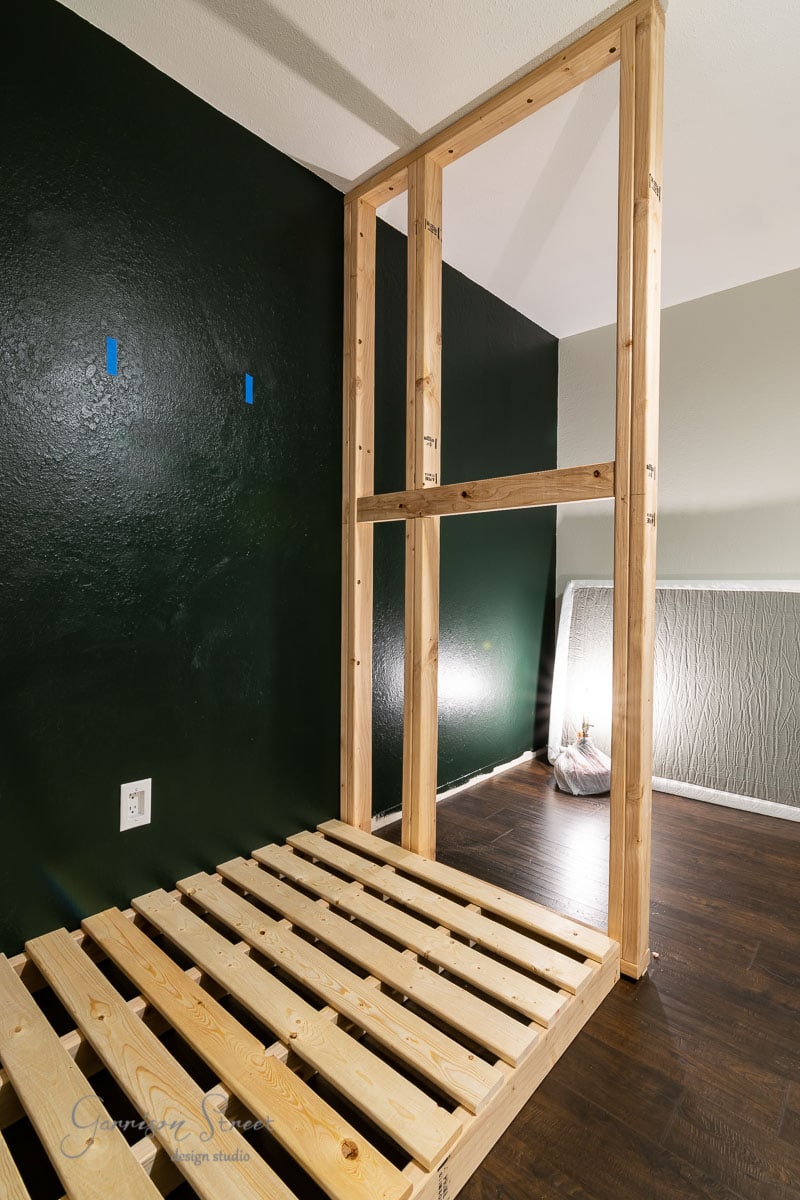 A wooden frame structure with a slatted base and built-in design stands in a room with dark green walls. Two blue tape markers adorn the wall, while a textured area with a lamp casts light and shadows across the floor.