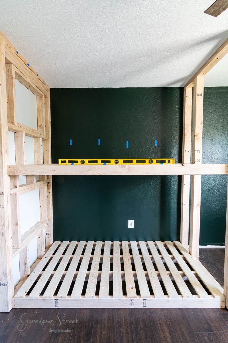 A wooden bed frame is under construction against a dark green wall. A yellow level tool rests horizontally on the frame, while blue tape marks measurements. The wooden floor adds warmth, and a single electrical outlet completes the scene.