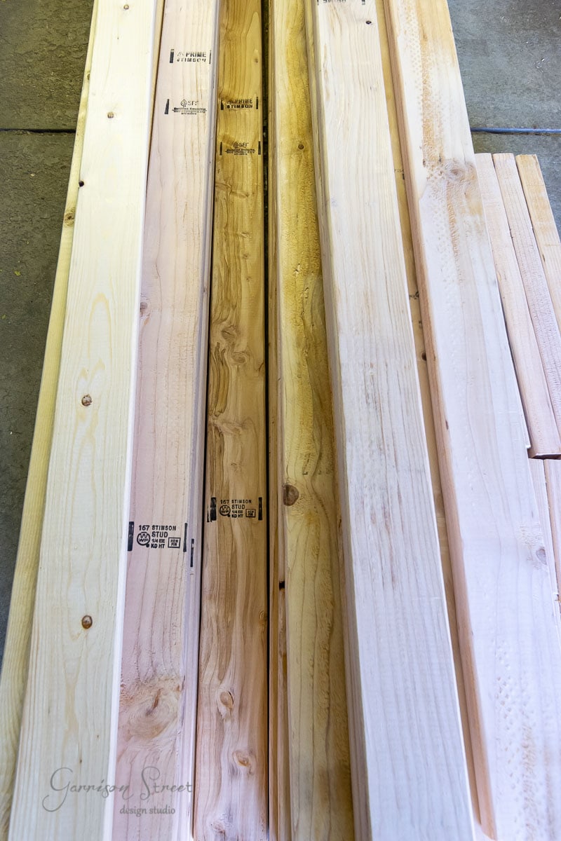 Stacks of long wooden planks are arranged side by side on a concrete floor. The planks showcase a light, natural wood finish with visible grain patterns and stamped text.