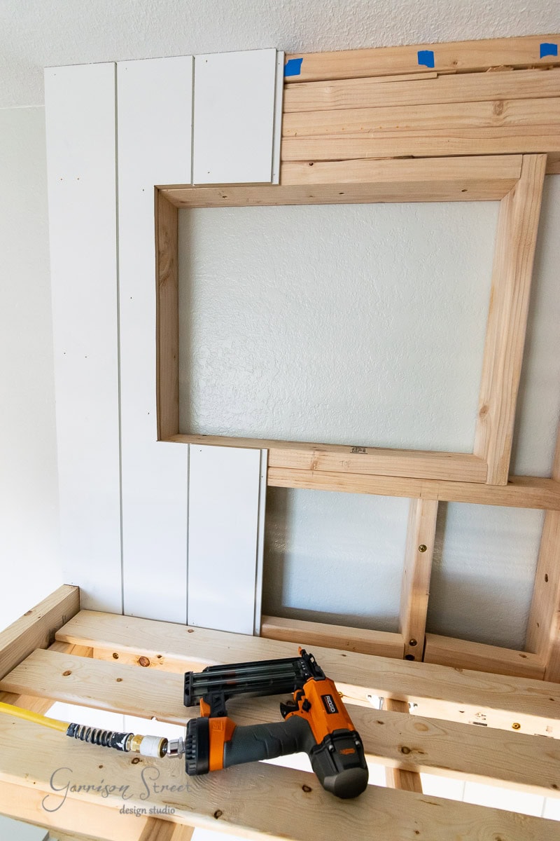 A partially constructed wood wall features a rectangular recess and painted white vertical panels. A nail gun, tape measure, and power drill sit on a wooden platform below. Painter's tape marks are visible on the wall above.