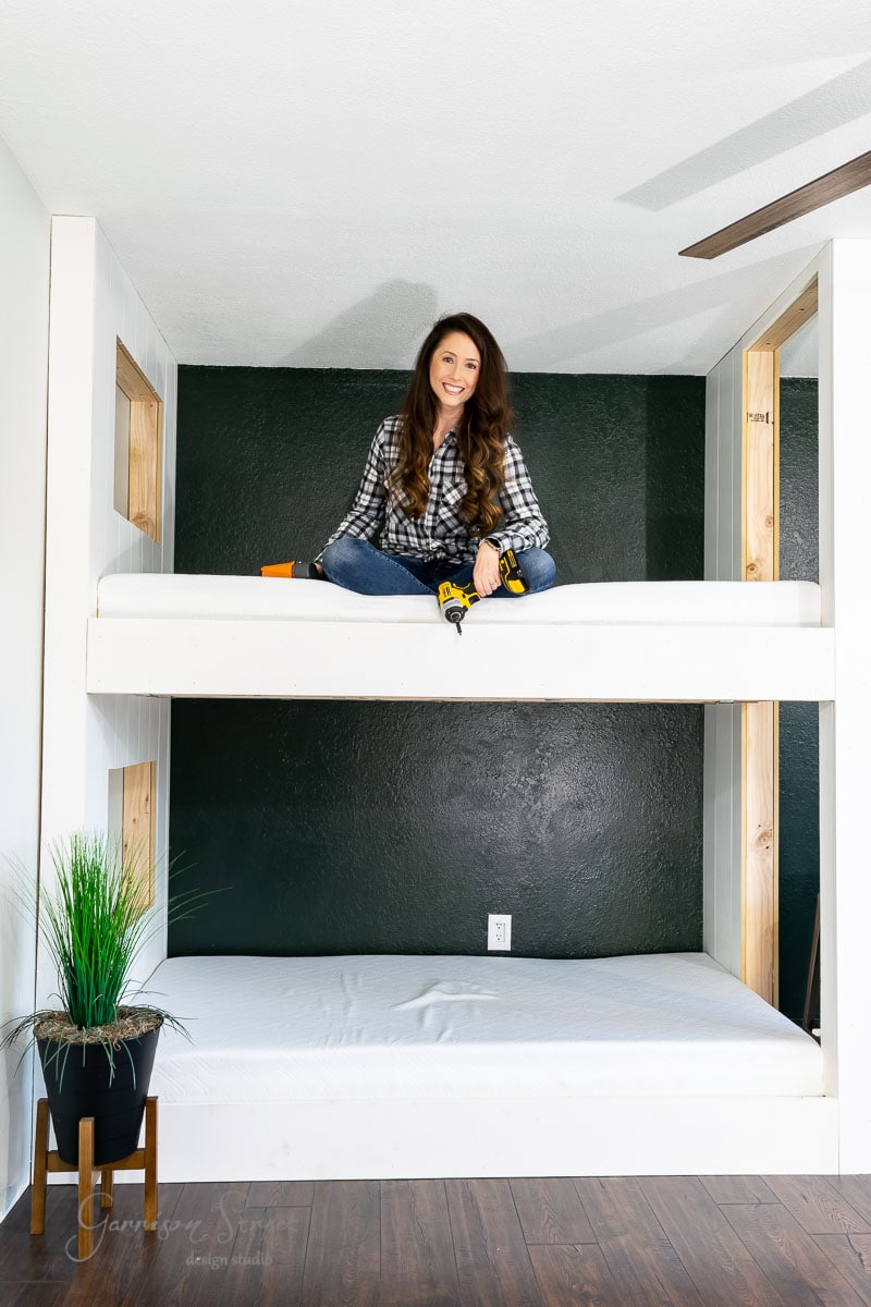A person in a plaid shirt and jeans sits on the top bunk of sleek, built-in bunk beds, holding a drill. A potted plant is on the wooden floor beside the bed, with a dark green wall as the backdrop.
