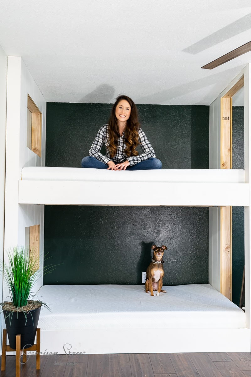 A woman sits cross-legged atop built-in bunk beds with a dark accent wall behind her. Below, a small dog relaxes on the bottom bed, adding charm. To the left, a potted plant completes the serene scene.
