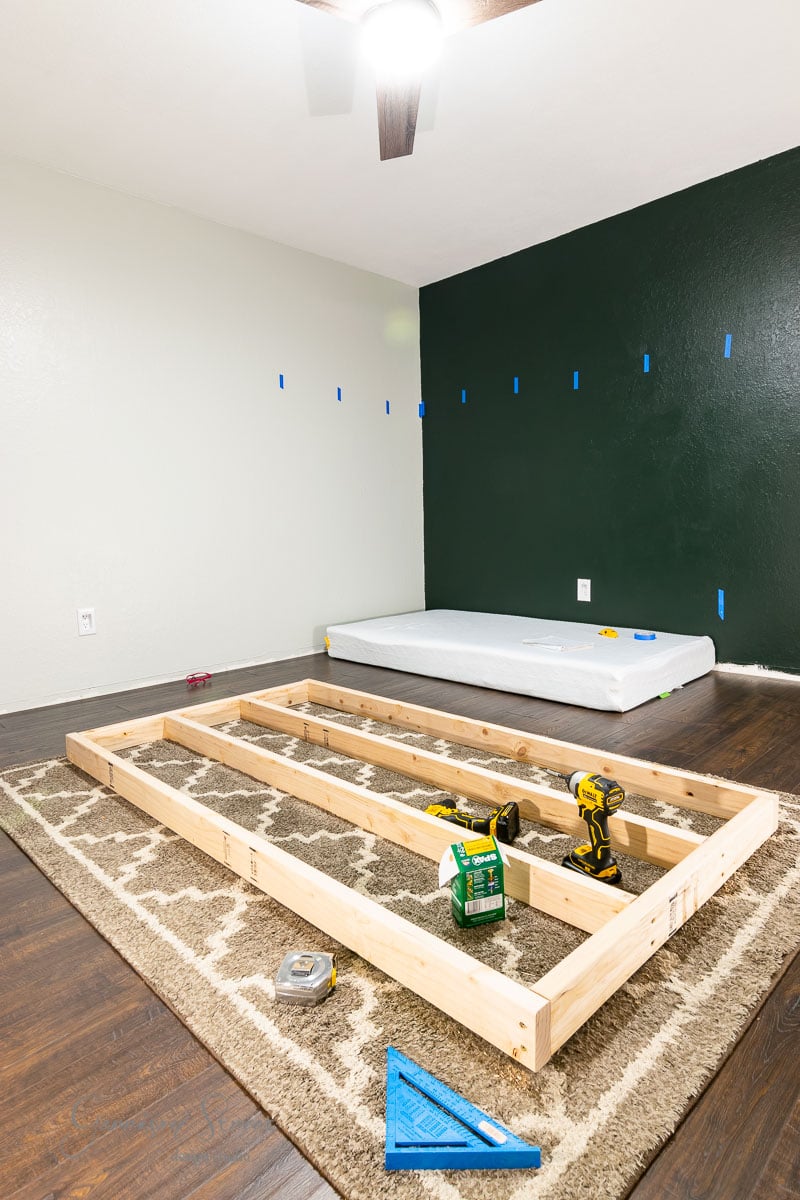 A partially assembled wooden bed frame sits on a patterned rug. A mattress leans against a green wall marked with blue tape. Tools like a power drill and square are scattered across the floor, all under a ceiling fan above.