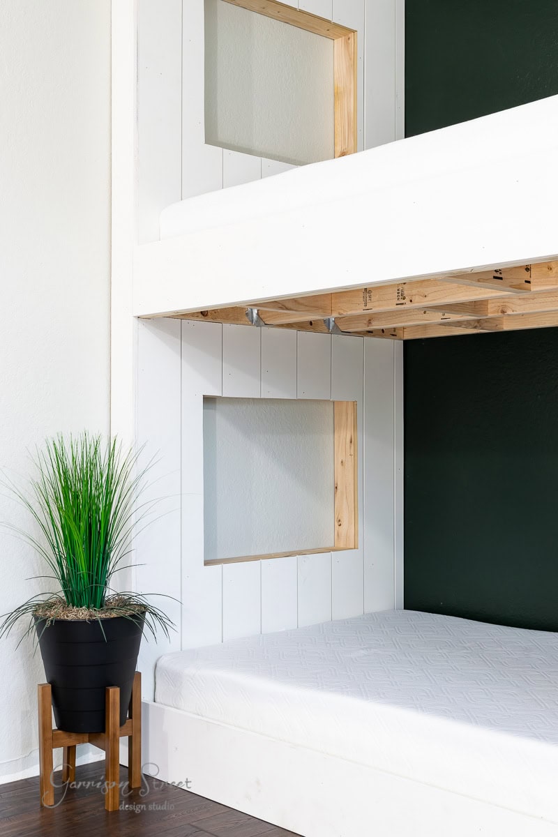 A modern bunk bed setup with white and wood accents against a dark green wall features sleek built-in design elements. A potted plant in a black and wooden stand on the wooden floor next to the bed adds a touch of greenery to the minimalist decor.