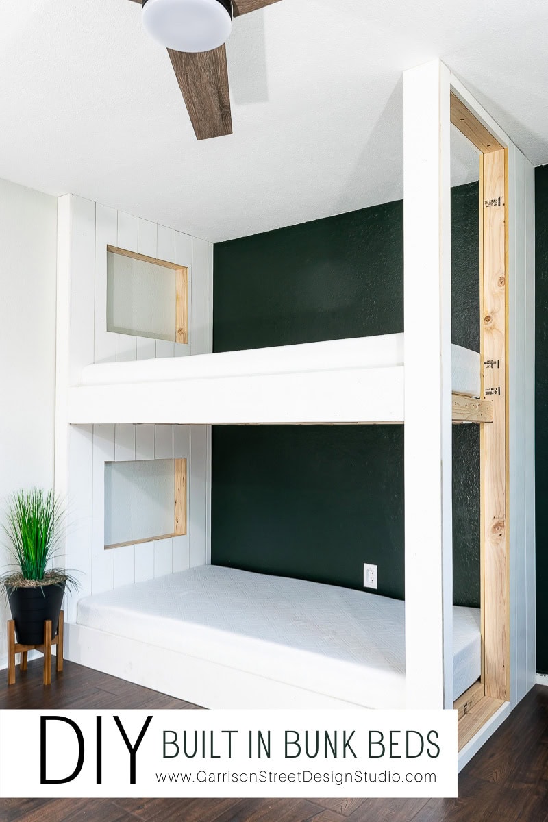Modern white and wood built-in bunk beds are seamlessly integrated into a wall with a dark green accent. A small potted plant sits on a nearby stool, adding a touch of nature. The bottom bunk is furnished with a mattress, while the top is empty.