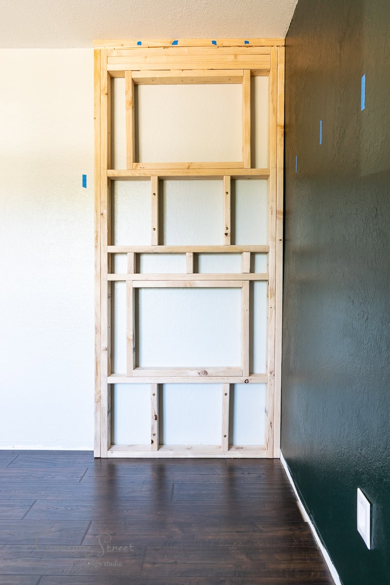 A partially constructed wooden frame for a built-in shelving unit stands against a wall. The hardwood floor and a dark accent wall complement the scene. Blue tape marks suggest measurements or alignment. The room is well-lit.