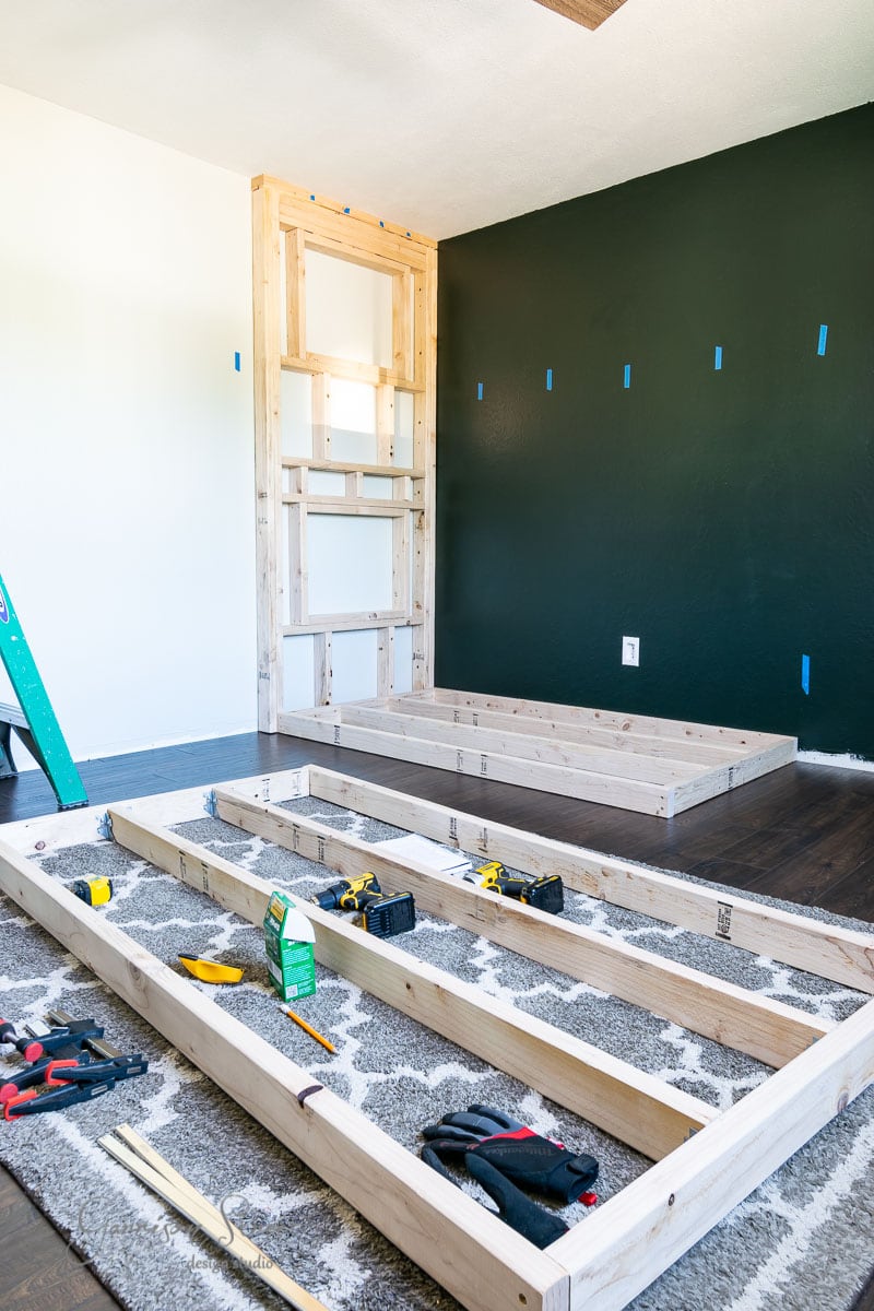 Wooden frame structures for a built-in shelving project are partially assembled in a room. Tools like drills and a tape measure are scattered on the rug-covered floor. A dark accent wall and a ladder are visible in the background.