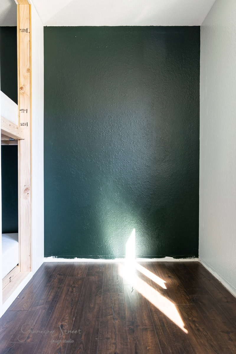 A small room featuring a dark green accent wall and a wooden bunk bed on the left, complemented by shiny hardwood floors. An IKEA Rast desk rests against the wall, while a bright triangle of sunlight on the floor offers a warm contrast to the deep wall color.