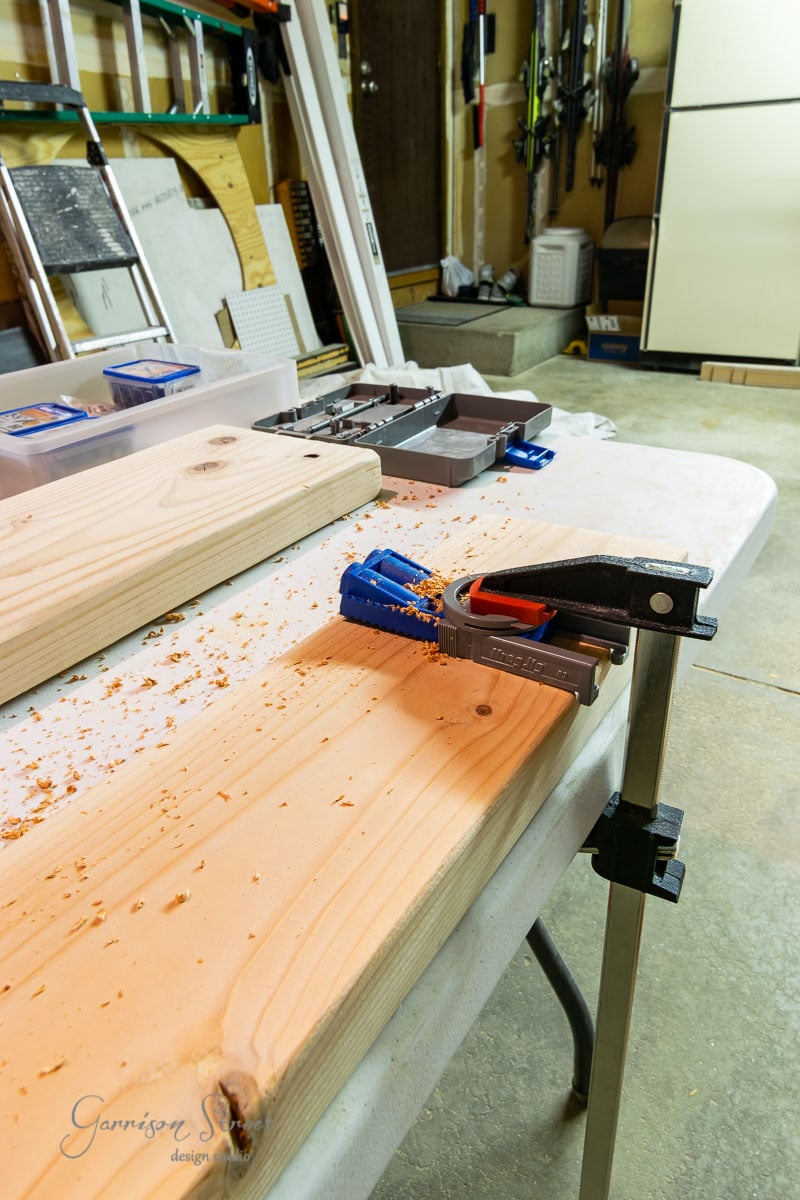 A workbench features a wooden board clamped for drilling. Surrounded by an array of tools and accessories, the table sits in a garage with various items neatly organized on shelves in the background.
