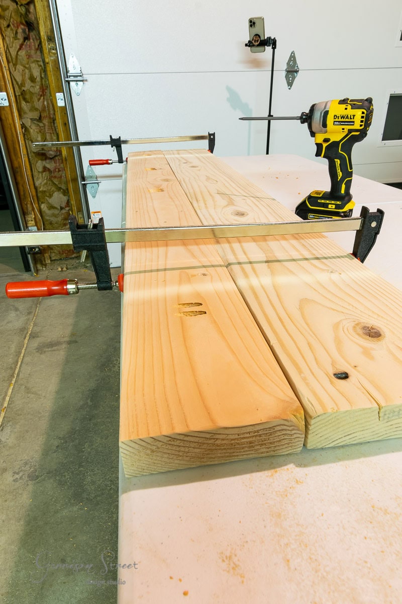 A workshop scene featuring two wooden boards clamped together on a sturdy desk. A yellow cordless drill rests on the boards, with a garage door and an IKEA Rast unit filled with various tools in the background.