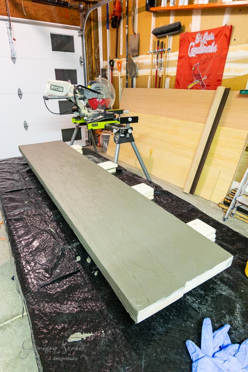 A long wooden board with a textured gray painted surface rests on sawhorses in a garage workshop. Power tools and a St. Louis Cardinals banner are also visible in the background.