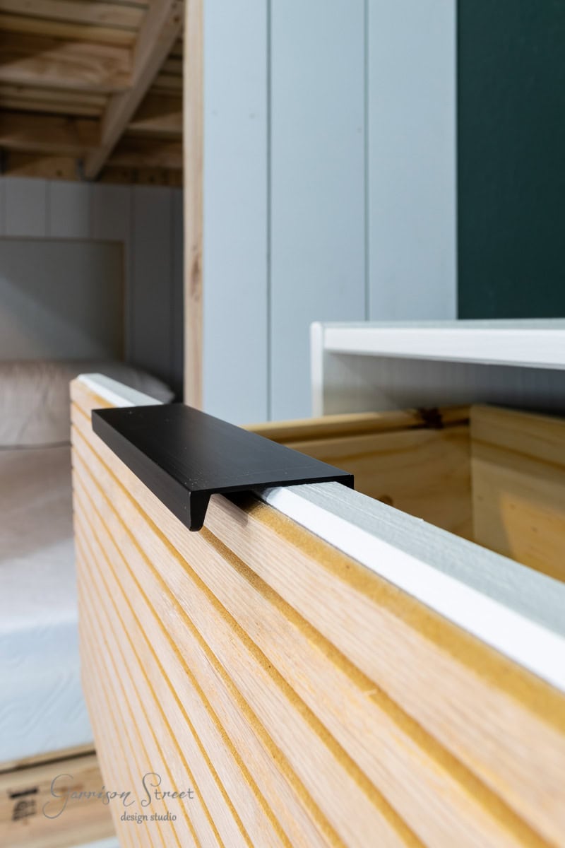 Close-up of a modern wooden bed frame with a slatted design. A black metal bracket accents the edge. The background reveals a glimpse of a room with wooden walls and another bed.