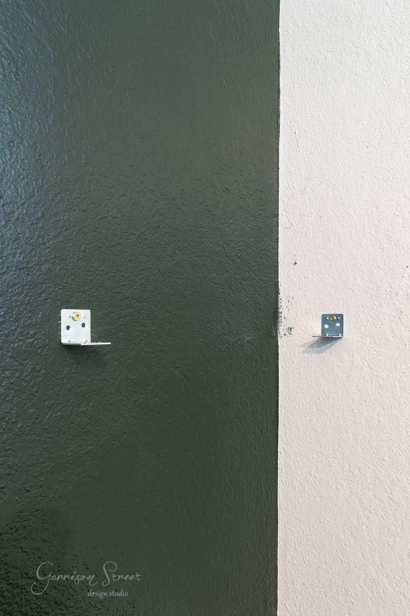 A close-up of a wall with two sections painted in dark green and light beige. Two small metal brackets are attached to the wall. The lower left corner features a watermark saying "Garrison Street design studio."
