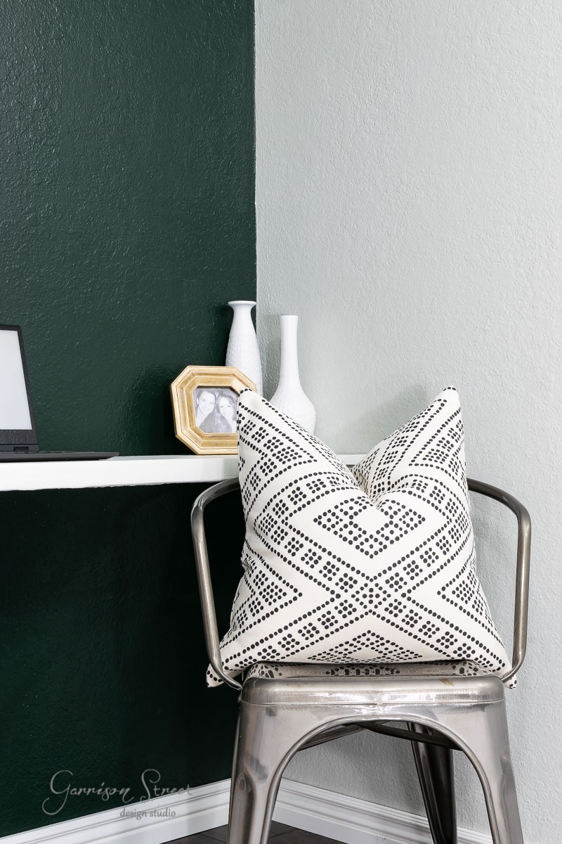 A metal chair with a black and white patterned cushion sits beside an IKEA Rast shelf. On the shelf are two white vases and a small photo frame. The wall is painted half white and half dark green, creating a striking backdrop for the minimalist desk area.