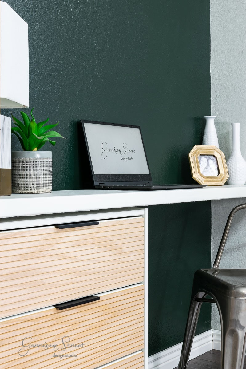 A modern office setup features an IKEA Rast wooden desk against a dark green wall. The desk holds a laptop, a small plant, a lamp, picture frame, and two vases. A sleek metal chair is tucked neatly under the desk.