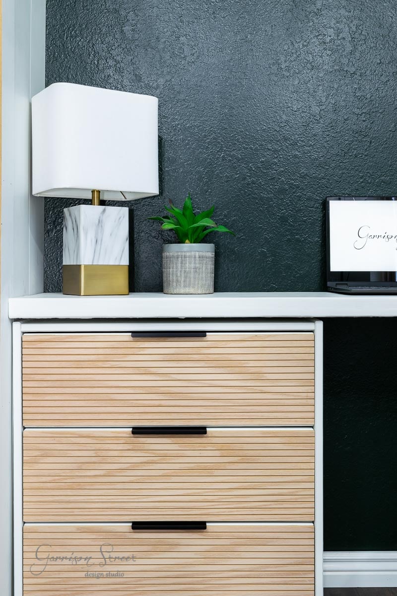 A modern setup features an IKEA Rast wooden desk with a small chest of drawers, a white and gold lamp with a marble base, a potted plant, and a square sign in cursive on a white background. The grey wall complements the sleek, white surface perfectly.