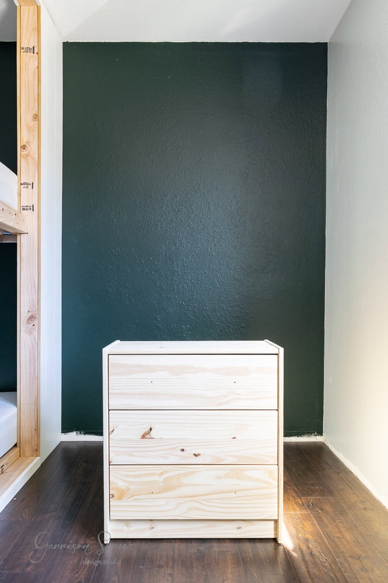 A simple, natural wood dresser like the IKEA Rast with three drawers stands on a wooden floor against a dark green wall. The room has a minimalistic and modern design with a cozy, warm ambiance.