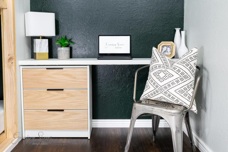 A modern home office space with a sleek IKEA Rast desk featuring a laptop, a small plant, and decorative items. A metal chair with a black and white patterned cushion sits nearby. The wall is painted dark green, contrasting beautifully with the light wooden flooring.