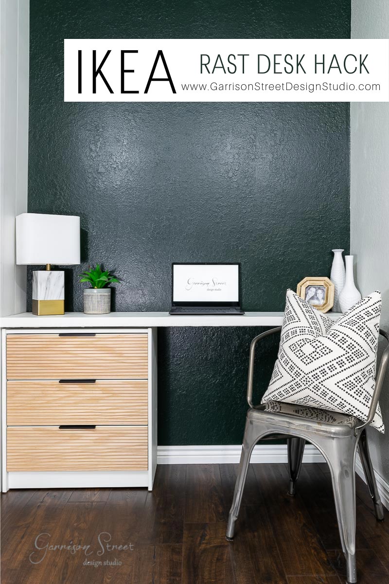 A modern home office features a DIY IKEA Rast desk with light wood drawers and a white surface. A lamp, plant, and framed artwork adorn the top. A metallic chair with a patterned cushion sits beside it. Dark green walls and wooden flooring complete the setting beautifully.