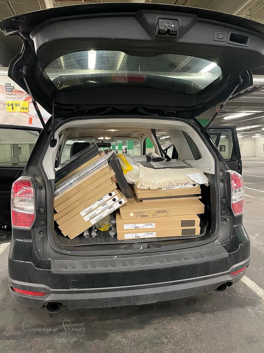 An SUV with the rear hatch open is filled with IKEA Rast flat-pack furniture boxes and a rolled carpet. It's parked in an indoor garage next to a pricing sign.