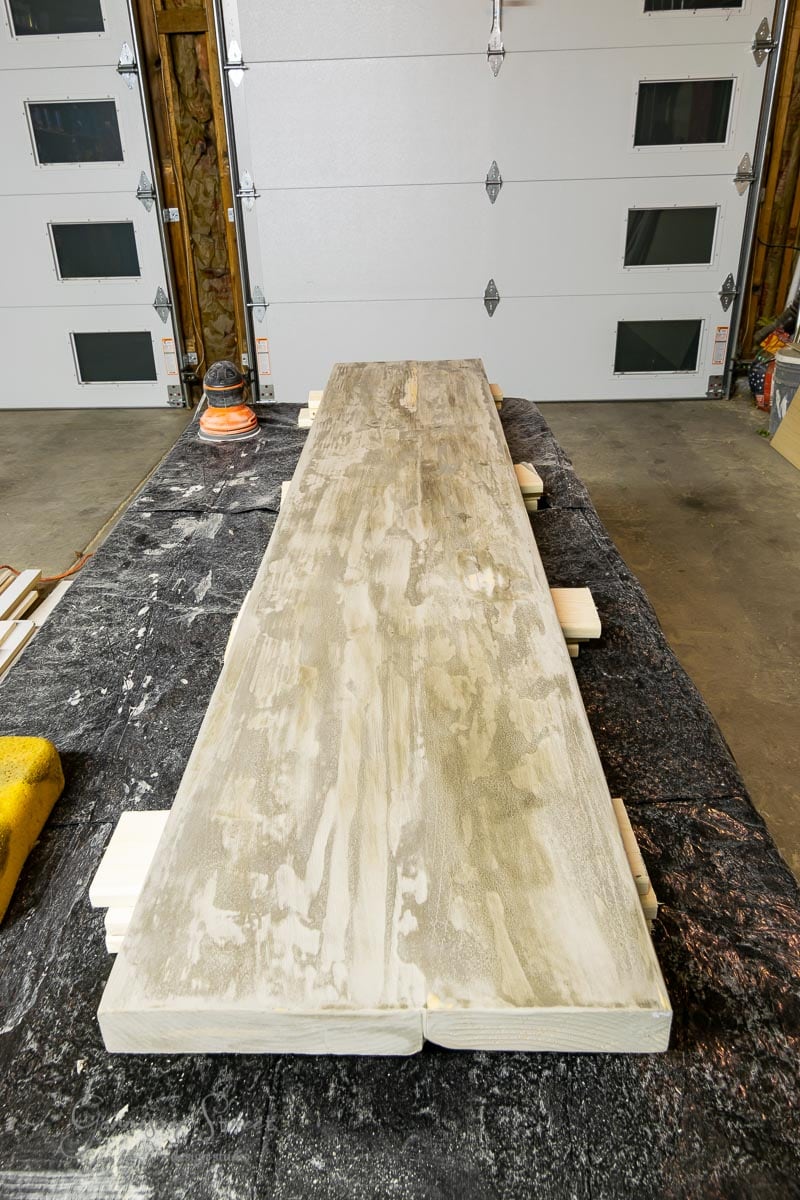 In a workshop, an IKEA Rast desk is being sanded. The long tabletop boasts a light wood color atop a black and white speckled surface, while an orange and black sander sits ready on the left edge of the table.