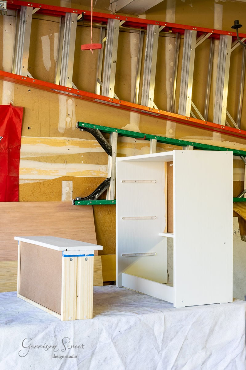 A garage workshop showcasing the assembly of a partially built white cabinet on a workbench. Various tools, alongside ladders, are neatly arranged on the wall in the background.