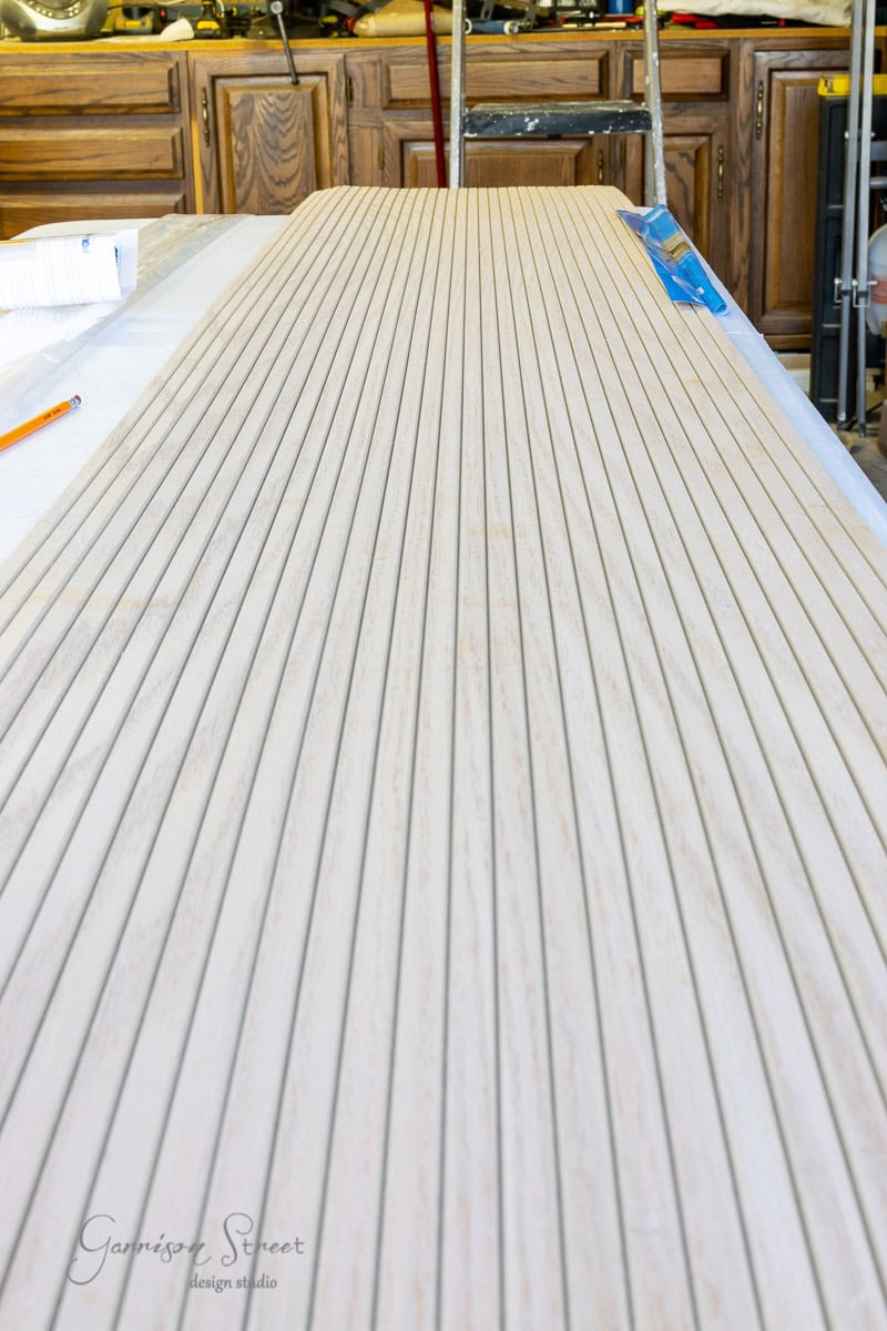 A long wooden tabletop with evenly spaced grooves sits on a workbench in a workshop. A pencil and ruler are nearby, suggesting ongoing measurement or construction work. Wood cabinets and tools are visible in the background.