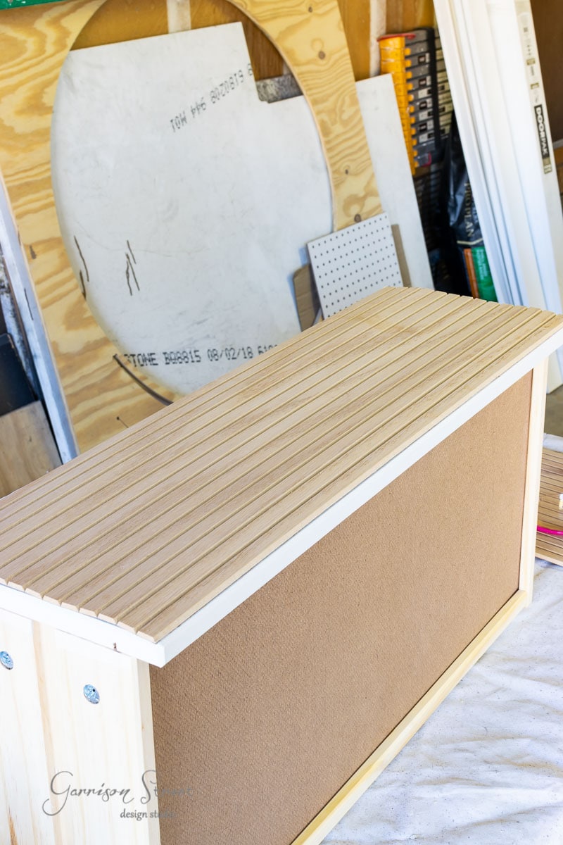 A DIY cabinet desk in progress features a plywood frame and a slatted wooden top. It stands surrounded by various tools and wooden planks in the vibrant atmosphere of a workshop.