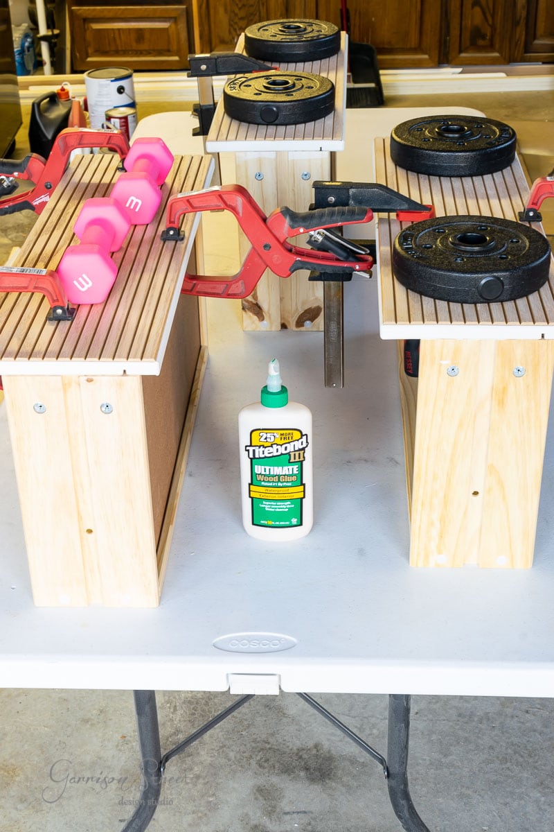 A woodworking project in progress features wooden boards clamped on a table. Various weights, including pink dumbbells, apply pressure. In the center, a bottle of "Titebond III Ultimate" wood glue ensures a sturdy build.