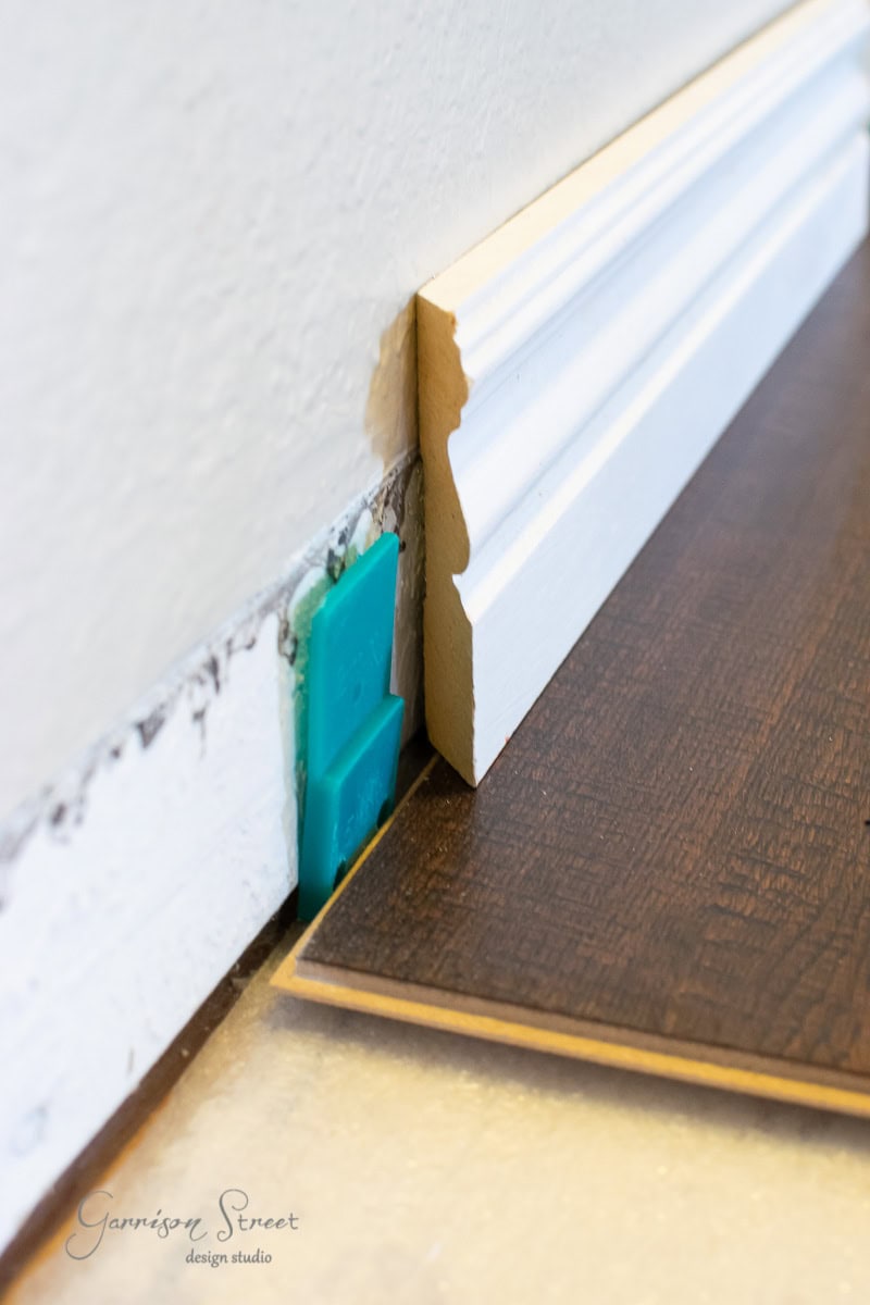 A close-up of laminate wood flooring installation in progress. A piece of dark wood laminate is being laid next to a white wall with baseboard. A blue plastic spacer is visible between the wall and the flooring. Image is credited to Garrison Street Design Studio.
