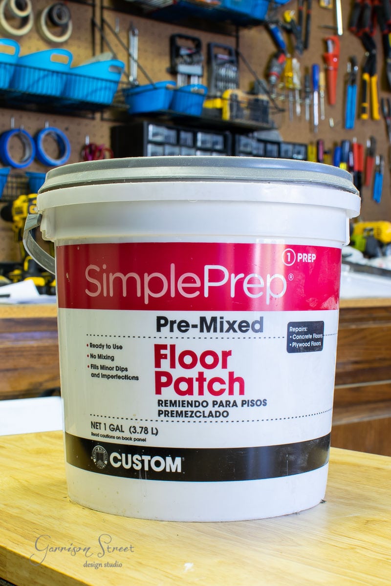 A bucket of SimplePrep Pre-Mixed Floor Patch sits on a wooden table, ready to tackle any project. In the background, a workshop showcases tools on a pegboard and shelves filled with essentials for installing laminate wood flooring. The label is bilingual, in English and Spanish.