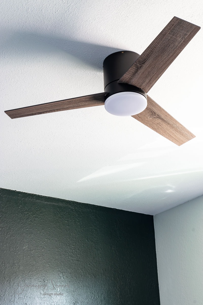 A modern ceiling fan with two wooden blades and a central light fixture is mounted on a white ceiling. The walls are painted green and white, creating a clean and minimalist look, beautifully complemented by the laminate wood flooring adding warmth to the space.