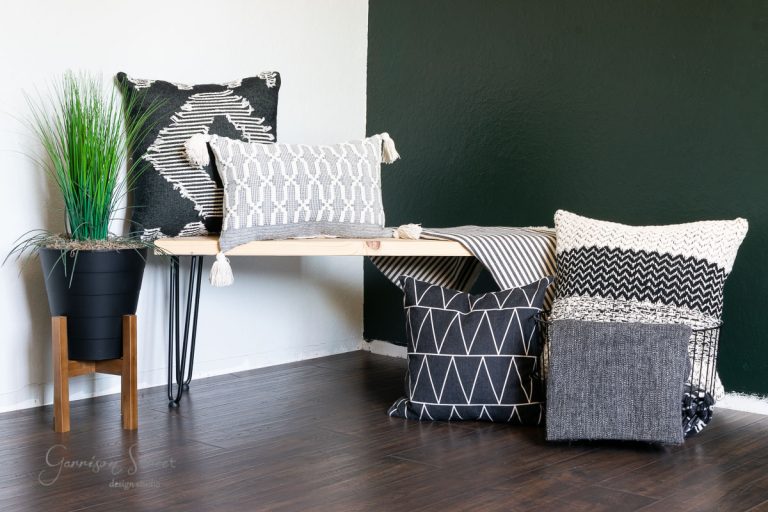 A modern room with a black and white theme features laminate wood flooring. A wooden bench holds patterned pillows in shades of black, white, and grey. A textured pouf sits beside a black plant pot with tall green grass. The dark wood floor complements the white and dark green walls beautifully.