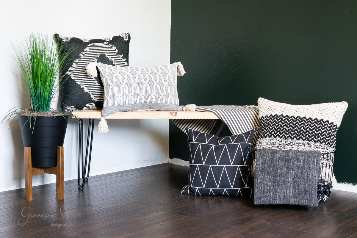 A modern room with a black and white theme features laminate wood flooring. A wooden bench holds patterned pillows in shades of black, white, and grey. A textured pouf sits beside a black plant pot with tall green grass. The dark wood floor complements the white and dark green walls beautifully.