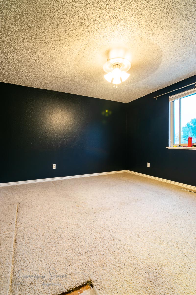 The empty room features dark blue walls and white carpet, though a patch is missing. A ceiling fan with a light casts a glow, complemented by natural light from a small window on the right. The windowsill holds a red object, contrasting against the suggestion of laminate wood flooring in the future.