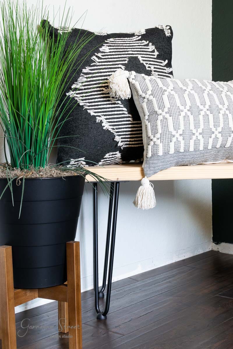 A modern interior scene showcases a wooden bench with black metal legs, adorned with two decorative cushions in black and white patterns. Beside the bench, a tall black planter holds green grass-like plants on sleek laminate wood flooring.