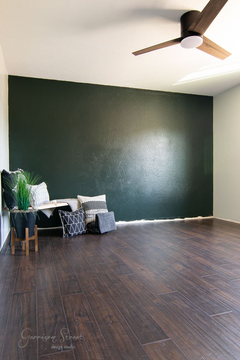 Minimalist room with a dark green accent wall and laminate wood flooring. A ceiling fan is above, with a cozy corner featuring a small bench, cushions, and a potted plant. Natural light illuminates the space.