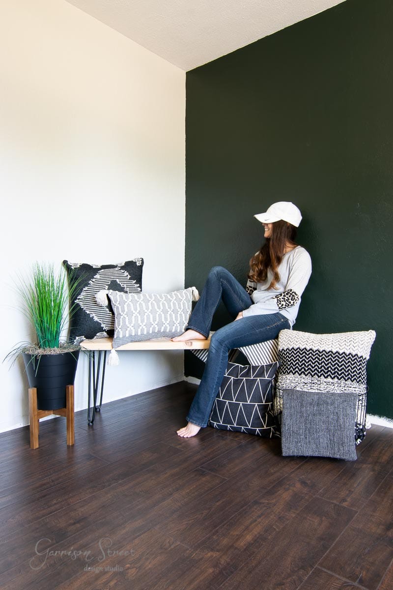 A person in a white cap and casual outfit sits on a wooden bench in a modern, minimalist room with laminate wood flooring. The room features a dark accent wall and several patterned pillows, while a small plant stands gracefully on the floor next to the bench.