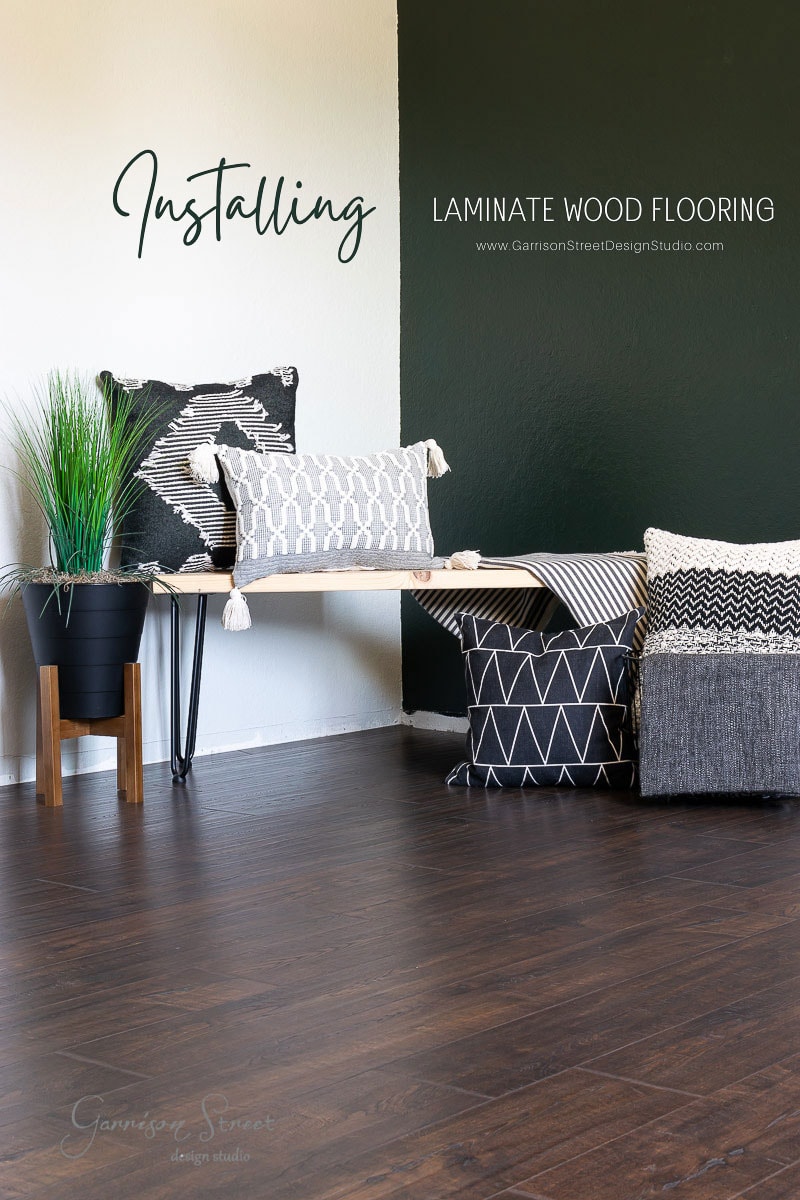 A cozy room features dark laminate wood flooring. Against a green and white wall, there's a wooden bench adorned with patterned cushions beside a small potted plant. Text on the wall highlights "Installing Laminate Wood Flooring" for an inviting touch.