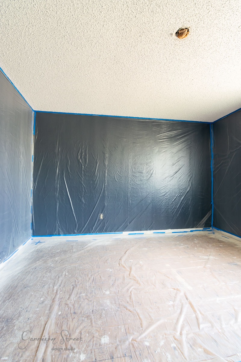 A room with dark painted walls and laminate wood flooring, protected by plastic covering parts of the ceiling. Blue painter's tape outlines the edges. The ceiling has a textured surface, and there's an uncovered electrical fixture hanging from above.
