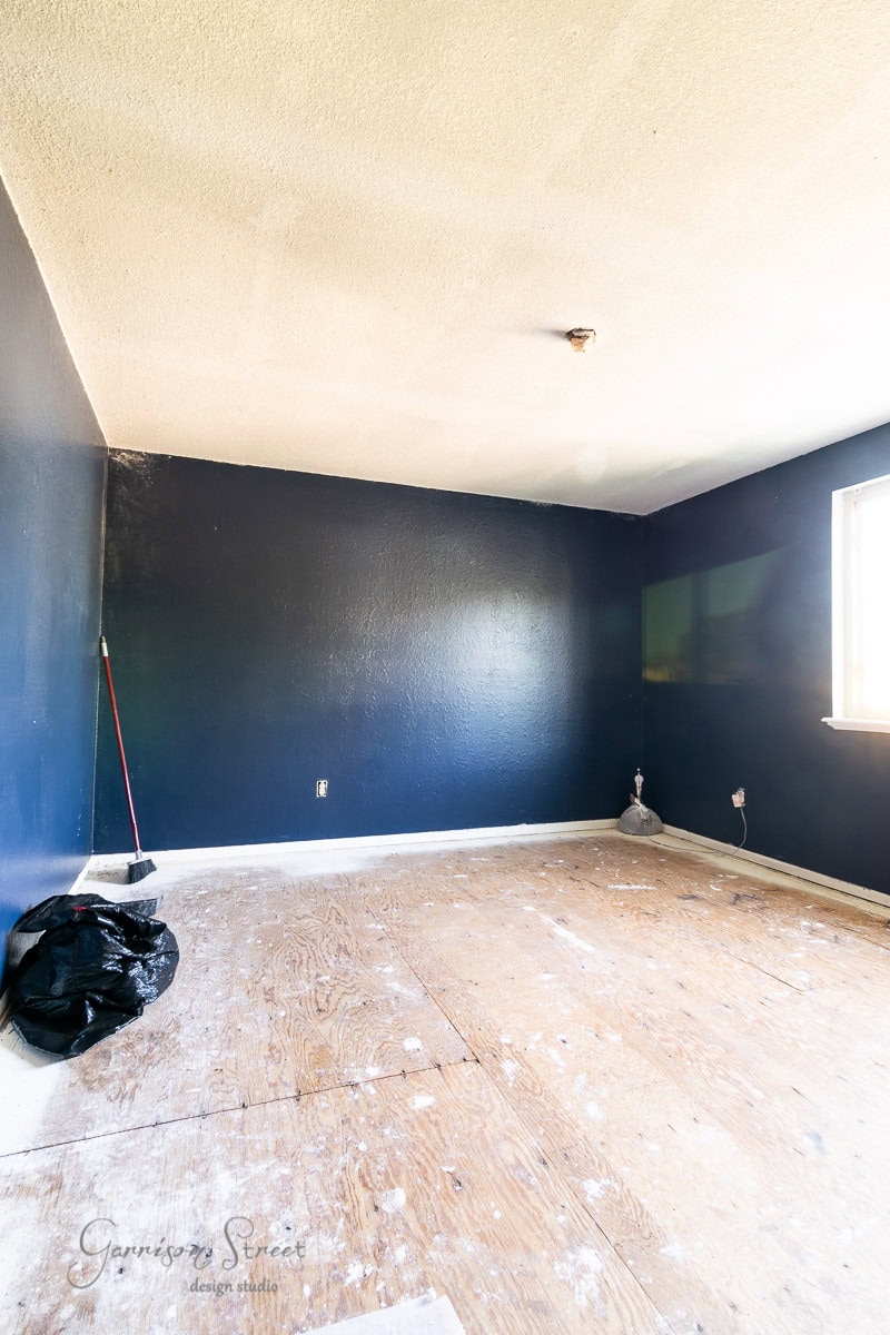 A bare room with dark blue walls and an unfinished wooden floor. A broom and a black trash bag are in the corner. Light streams through a window on the right, highlighting the simple, empty space, envisioning the potential transformation with laminate wood flooring.