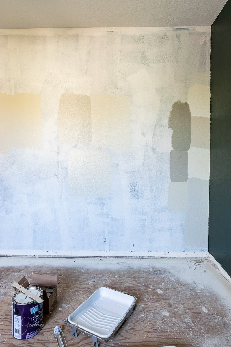Paint swatches adorn a partially painted wall, displaying various shades of white and gray. A dark green wall contrasts on the right. The unfinished floor awaits its future laminate wood flooring, accompanied by a paint tray and paint cans scattered in the foreground.