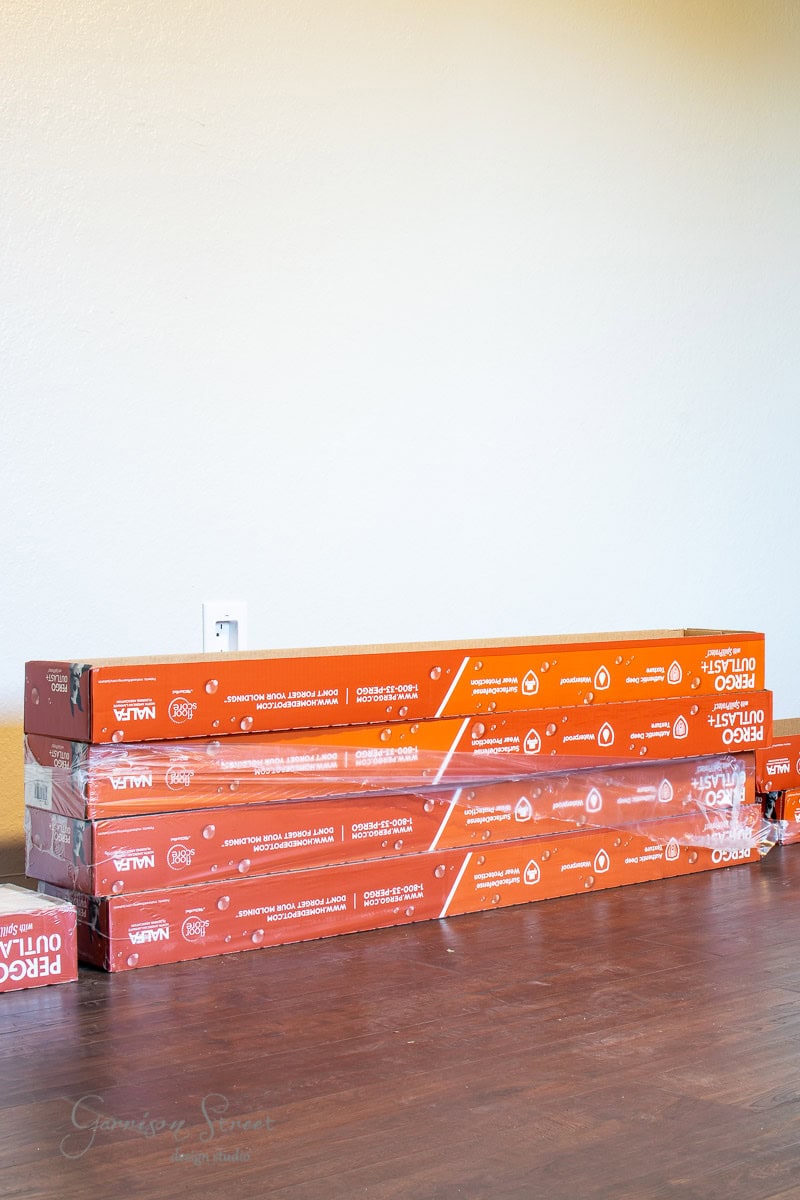 Four stacked boxes of Pergo Outlast+ laminate wood flooring rest against a wall. The orange boxes, featuring white text and icons, complement the room's wooden floor beautifully.