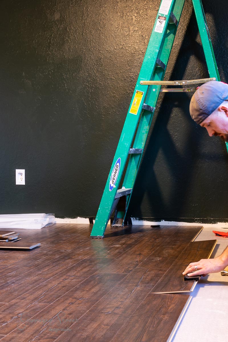 A person is installing laminate wood flooring in a room with dark green walls. A green ladder leans against the wall, and there is an electrical outlet nearby. The person is kneeling and using tools to align the panels meticulously.