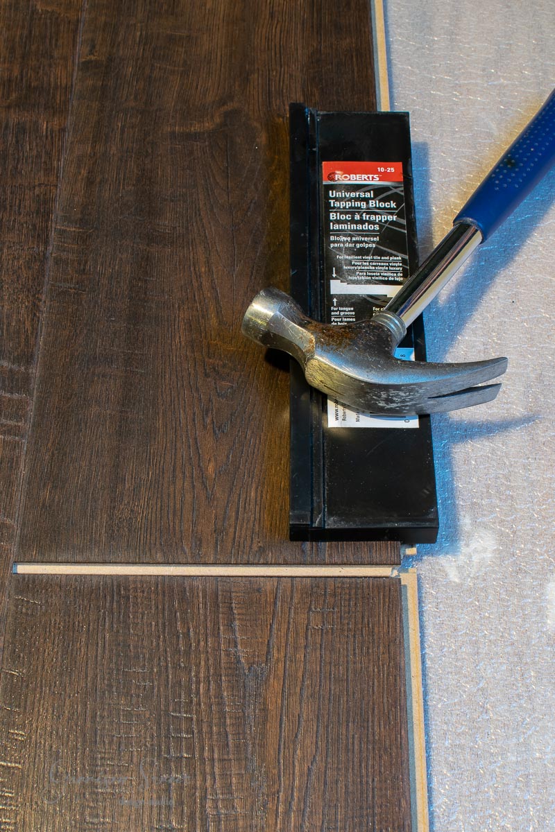 A hammer rests on a pack of flooring installation tools, with a plank of dark laminate wood flooring partially laid on a silver underlayment. A small gap is visible between the end of the plank and the adjacent one.