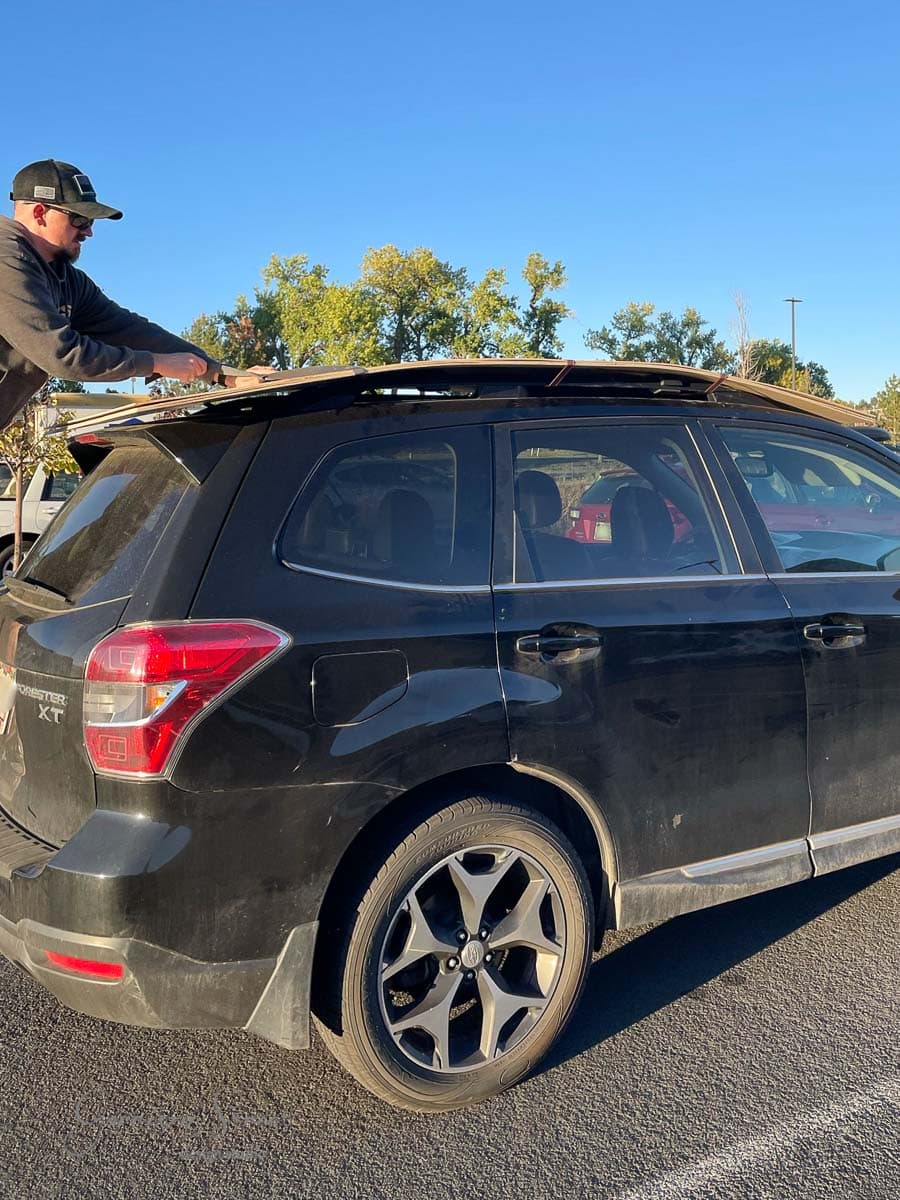 A person secures a large wooden closet door on the roof rack of a black SUV. The vehicle is parked outdoors on a sunny day, sliding smoothly into place with green trees in the background.